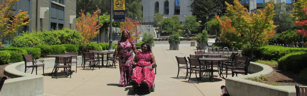 Two African women, one a power wheelchair user, in a courtyard.