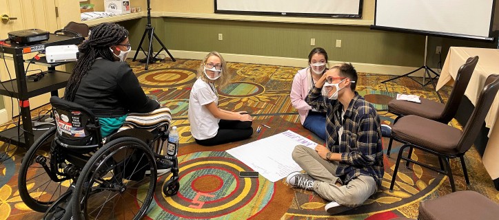 Group of students sitting on the floor listening to woman in a wheelchair. Students wearing clearing masks.
