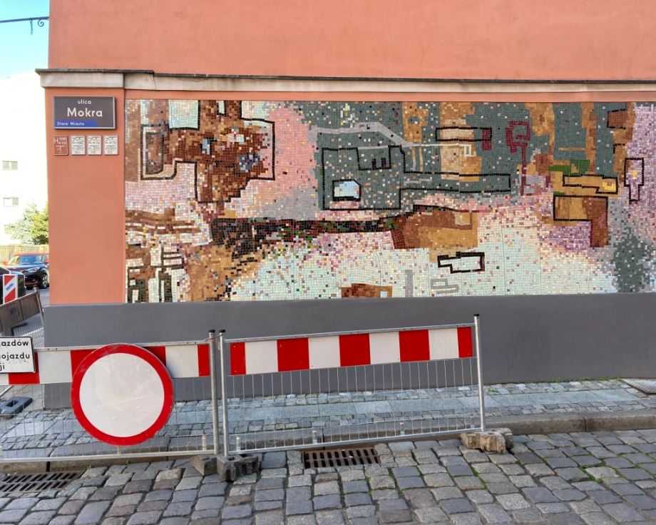 On a cobblestone street, a metal fence creates a barrier in front of a coral-colored building whose wall is covered with a large abstract mosaic mural in pastel and neutral tones