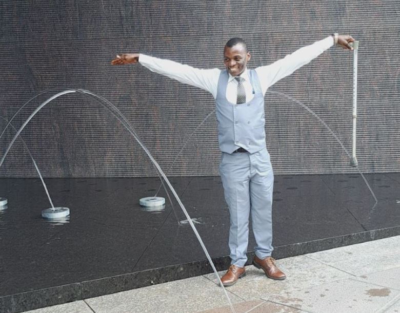 Haunah, dressed in blue and white suit with arms outstretched in front of a fountain