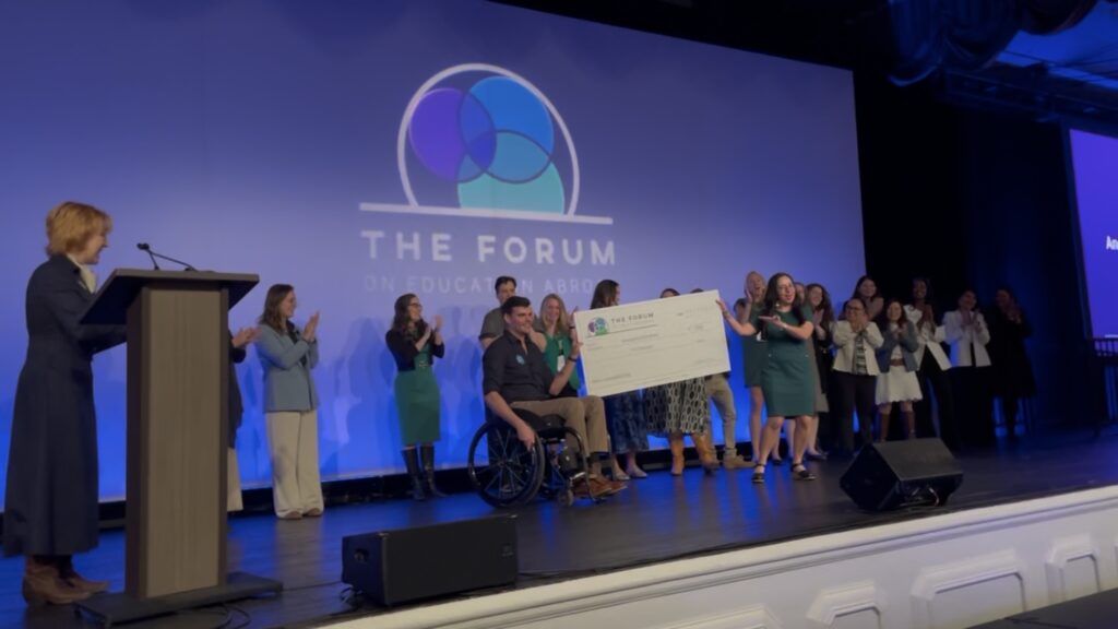 Kevin Sullivan, who uses a wheelchair, on stage with a dozen other people who are cheering for him, accepting large check from a person. Behind them is a wall with the Forum logo.