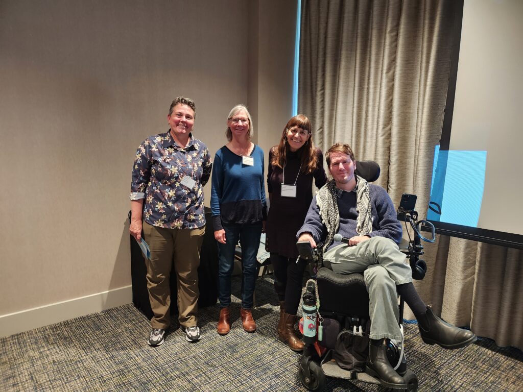 Four people gather together in a conference room, smiling at camera. They wear business casual clothing and conference badges. Three people stand and one person sits in a power wheelchair
