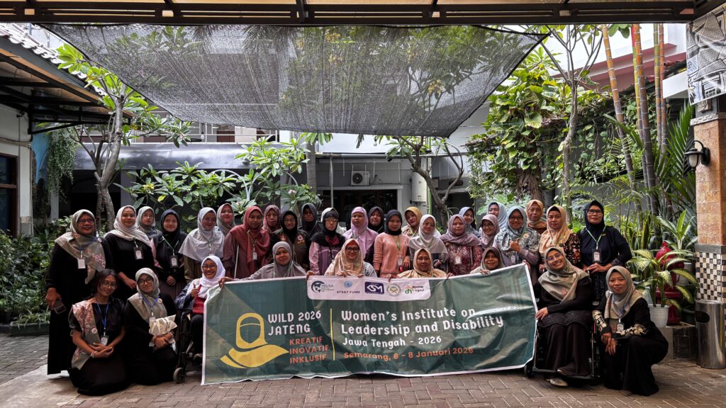 A group of thirty-three women of different ages with different disabilities are gathered in a plant-filled courtyard. They are holding a large green banner that says "Women's Institute on Leadership and Disability Jawa Tengah - 2026".