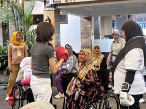 A smiling woman in a wheelchair wearing black boxing gloves is preparing to punch the gloved hands of the self-defense and Muay Thai coach. Six other women with disabilities sit or stand around them to cheer her on and wait for their turns.
