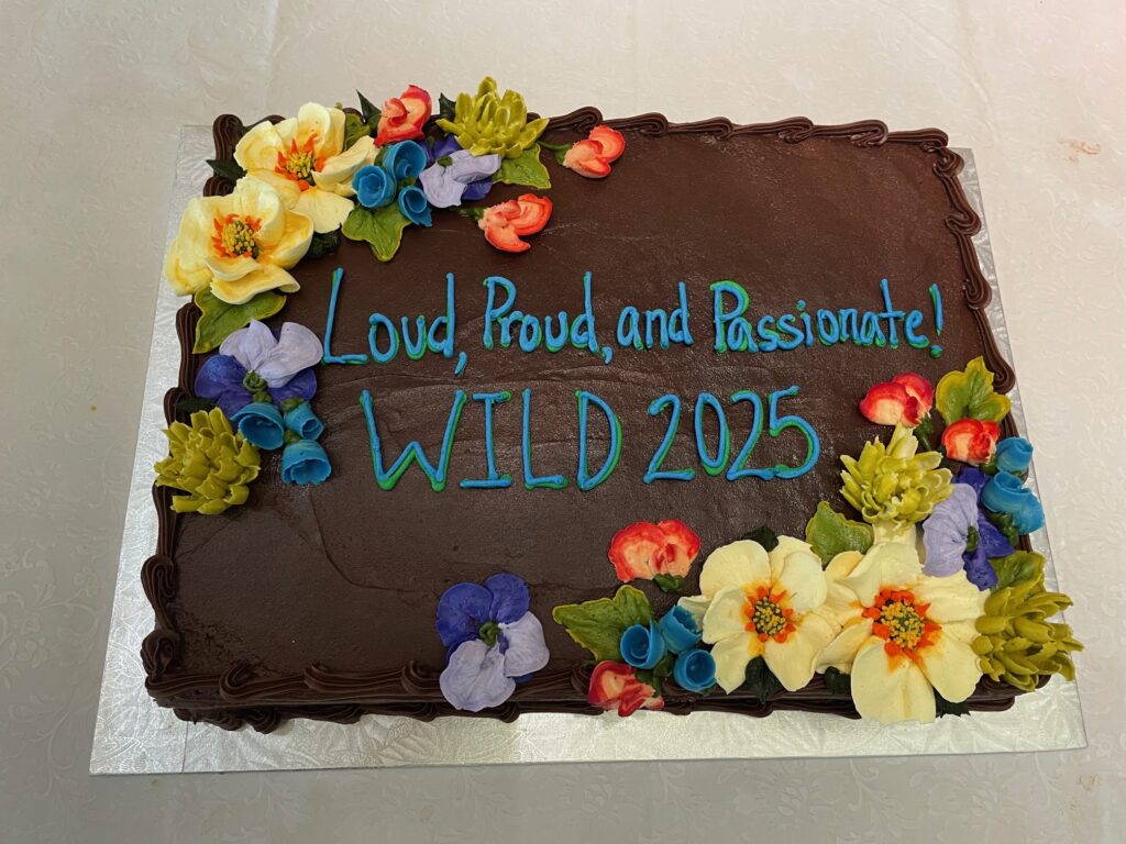 Sheet cake frosted with chocolate icing, colorful icing flowers, and written in frosting: Loud, Proud and Passionate! WILD 2025!"