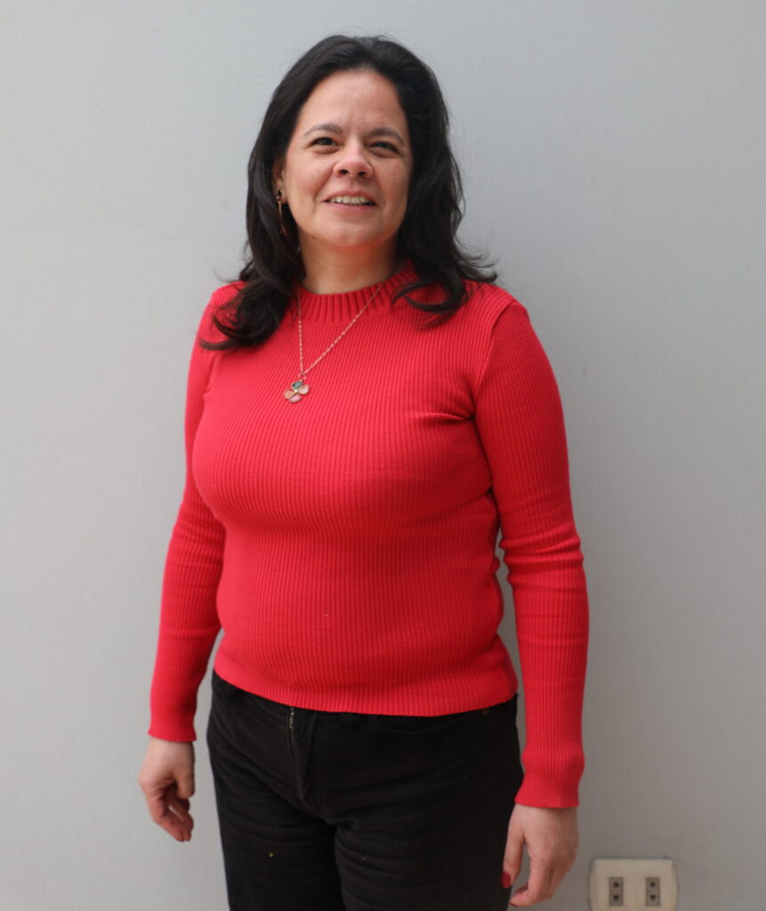 Angela stands against grey background wearing red long sleeve shirt as she smiles at the camera