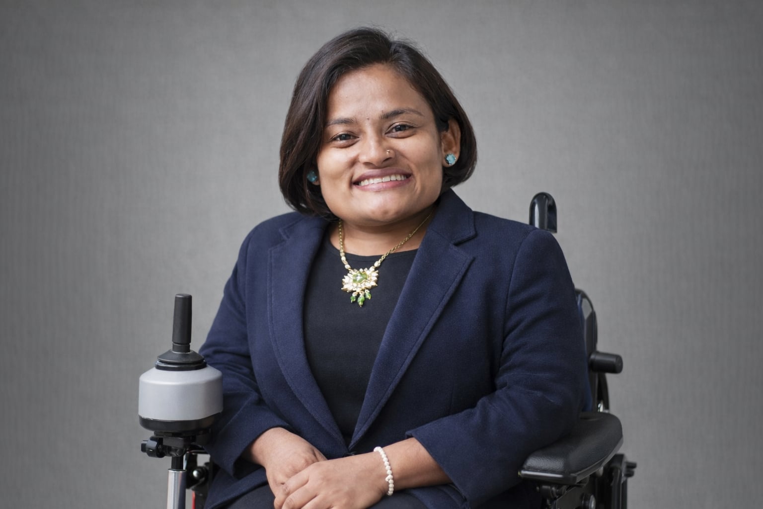 Anjana smiles at the camera, wearing dark blue blazer, seated in power wheelchair