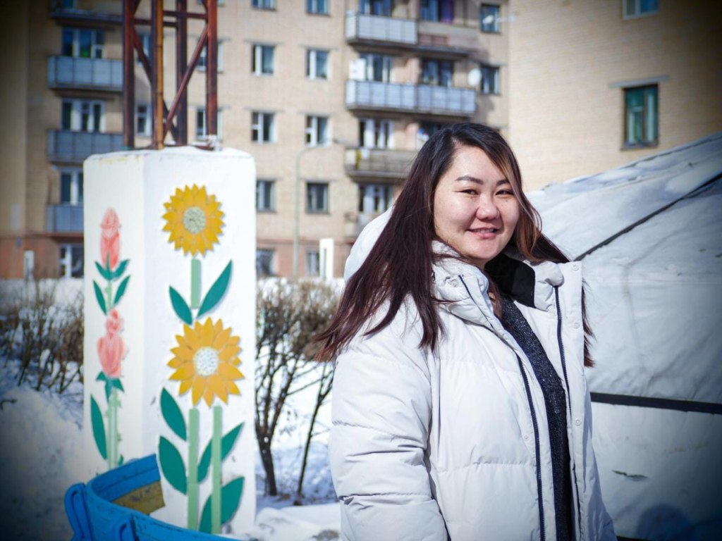 Dulya stands outdoors with buildings and a mural behind her. She wears large white winter jacket.