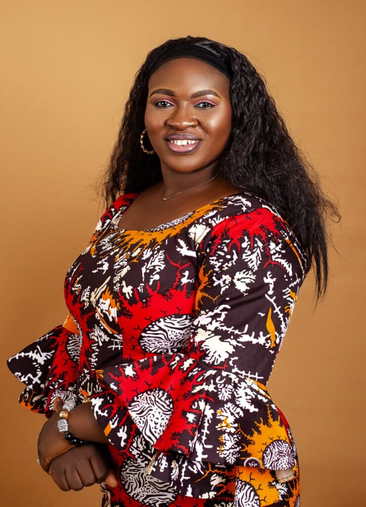 Hellen stands with hands folded in front of her in bold red and white print dress; she looks over her shoulder to smile brightly at the camera