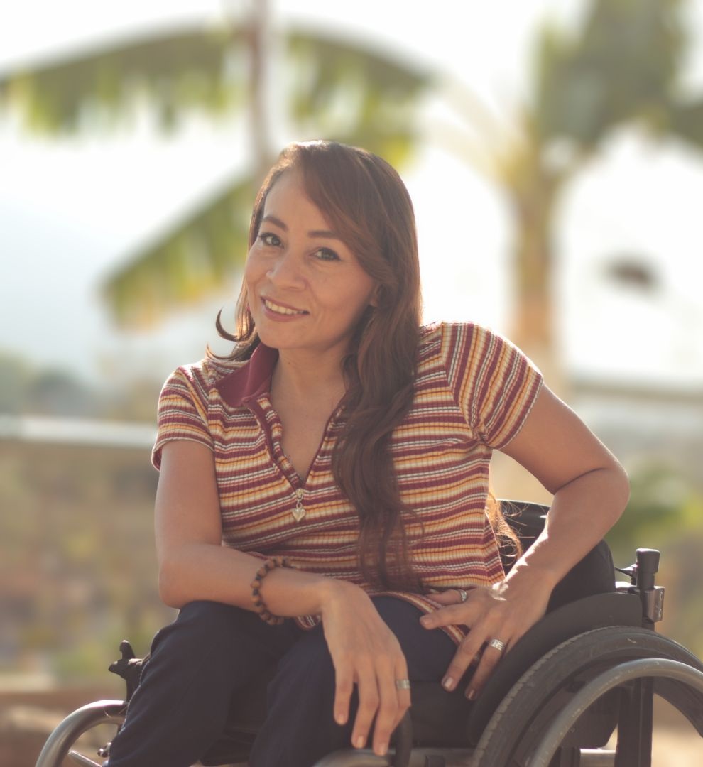 Jenny leans forward in her wheelchair, smiling at camera with trees in background