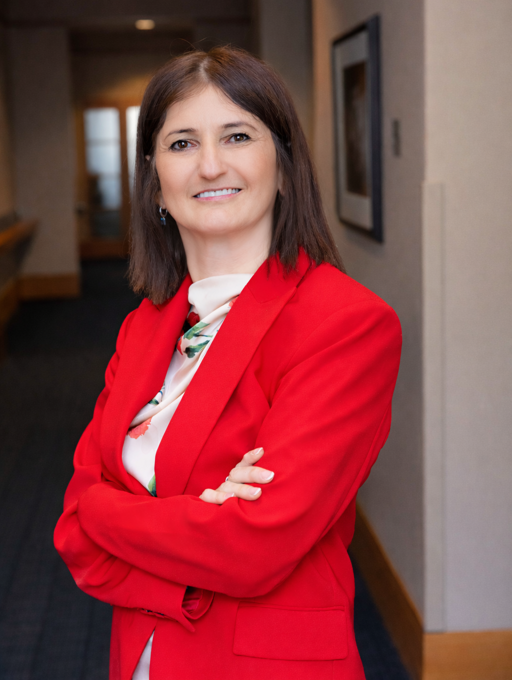 Karine in bright red suit, with arms crossed, gazing toward the camera standing in a hallway
