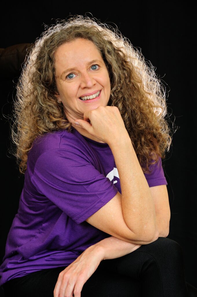 Mila portrait, resting chin on hand, smiling at camera, wearing purple shirt against black background