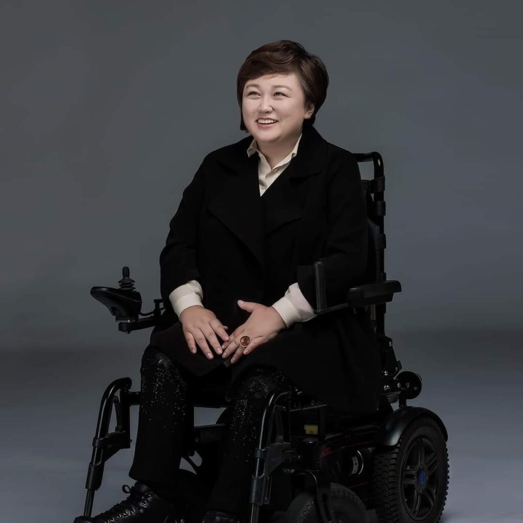 Miyeon seated in power wheelchair wearing dark colors against dark grey background, smiling away from camera