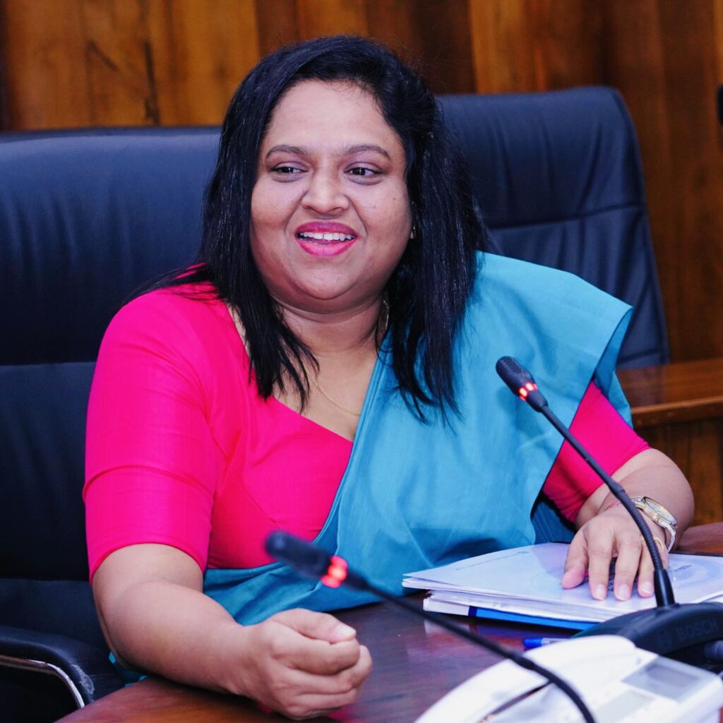 Rasanjali sits at a desk equipped with microphone. She is seated in a large leather chair, and wears a blue sash over hot pink dress.