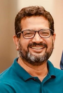 Shafiq portrait wearing teal polo shirt, smiling at camera wearing glasses