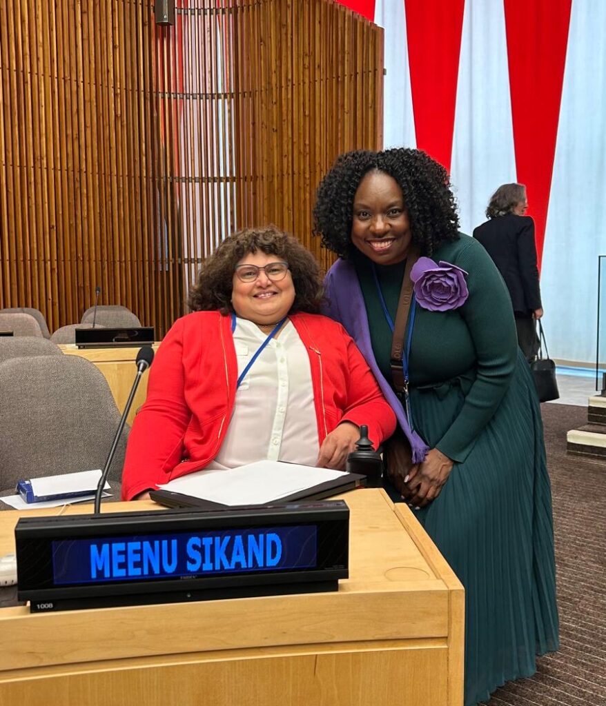Meenu Sikand sits behind a speaker podium with her nameplate, with her colleague beside her.
